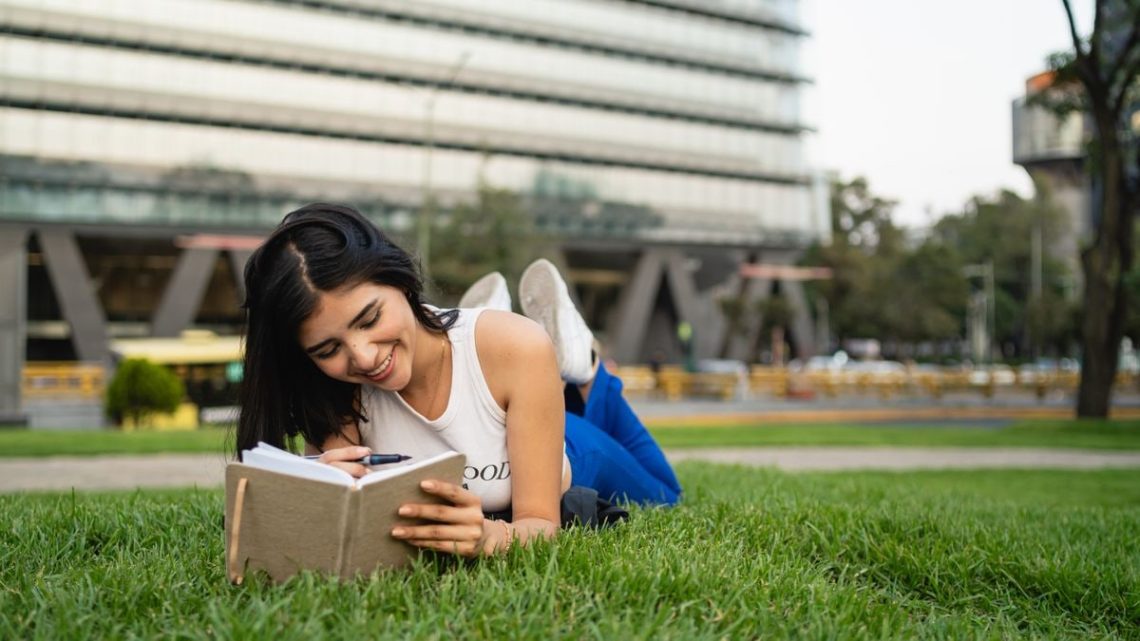 Smiling woman lying on grass reading a notebook in a city park on a sunny day.