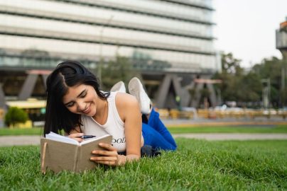 Smiling woman lying on grass reading a notebook in a city park on a sunny day.