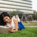 Smiling woman lying on grass reading a notebook in a city park on a sunny day.